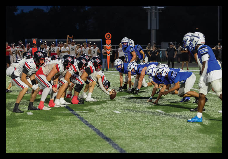 Two opposing football teams in helmets and uniforms are lined up facing each other on a field, ready for the next play during a night game. Description generated by AI