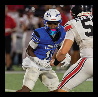 A person wearing a blue sports jersey and white padded pants is running on a grassy field during a game. Description generated by AI