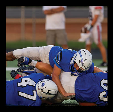 Three football players in blue jerseys and white helmets are on the ground during a game, with one player tackling another. Description generated by AI