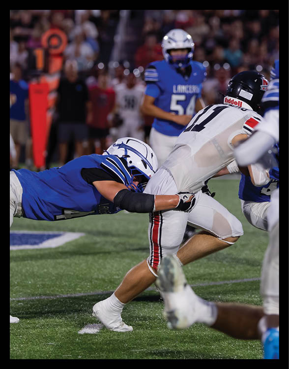 A person in blue sports gear and helmet is diving forward to tackle another person wearing white pants with red and black stripes during a football game on a turf field. Description generated by AI