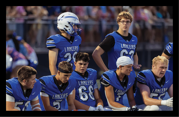 A group of people wearing blue football uniforms with “Lancers" on the front are sitting and standing on the sidelines during a game. Description generated by AI