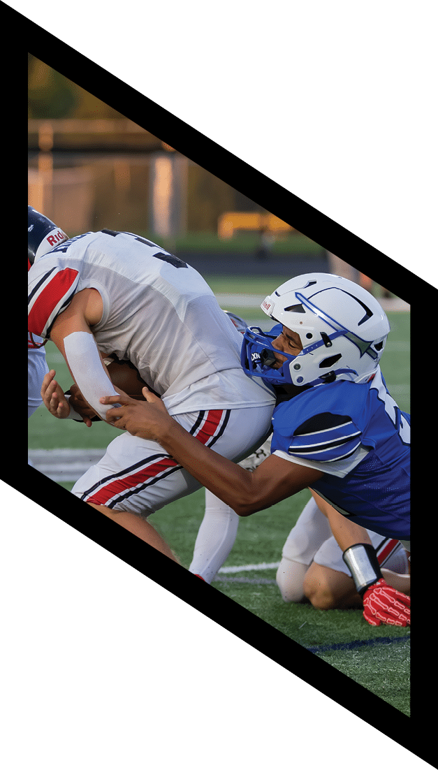 Three people wearing football uniforms are engaged in a play on a field, with one person in white being tackled by another in blue and a third person in white nearby. Description generated by AI