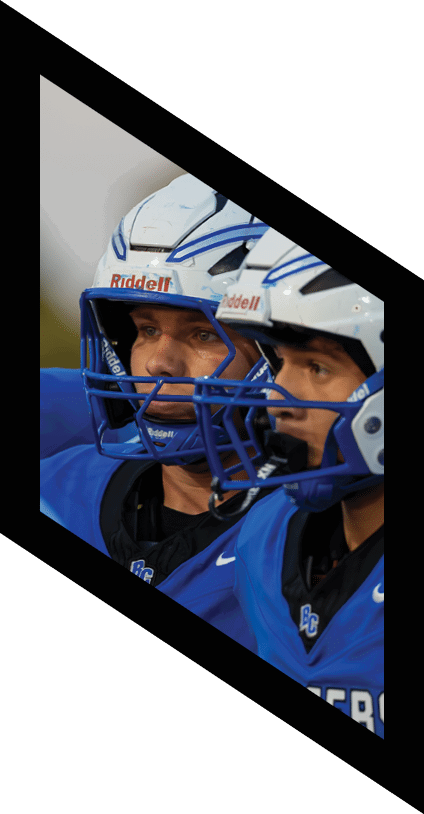 Two people wearing blue football helmets and blue jerseys are engaged in a close interaction during a game. Description generated by AI