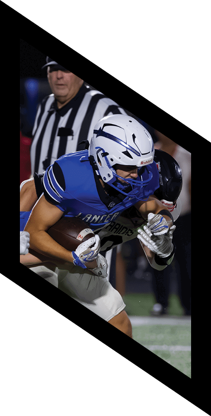 A person wearing a blue football jersey and white helmet is holding a football tightly while being tackled by another person in a black helmet and gray uniform on a football field with a referee in the background. Description generated by AI