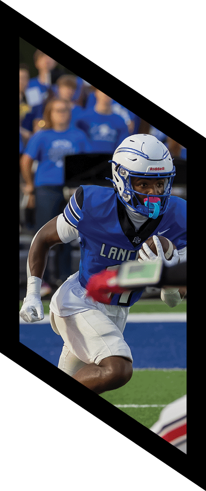 A person wearing a blue football uniform with the word “Lancers" is running with a football while another player in a white and red uniform reaches out toward them on a football field. Description generated by AI