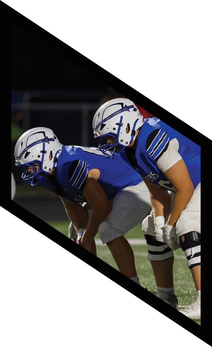A group of people wearing blue sports uniforms and white helmets are lined up on a football field preparing for a play. Description generated by AI
