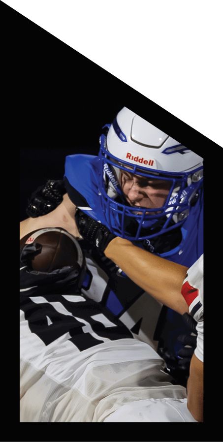 People wearing football uniforms are engaged in a tackle during a nighttime game on a grass field. Description generated by AI