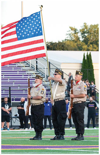 Three people in uniform stand on a football field holding rifles while one salutes beneath a large American flag with spectators in the background. Description generated by AI
