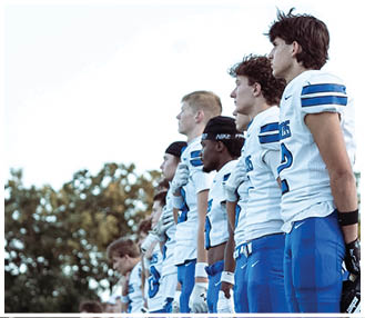 A line of adults wearing blue and white football uniforms stand side by side holding their helmets in front of them outdoors. Description generated by AI