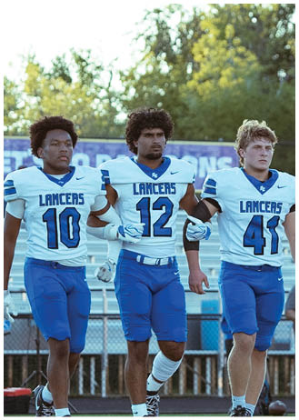 Three people in blue and white football uniforms with the word LANCERS walk arm in arm on a sports field with trees in the background. Description generated by AI