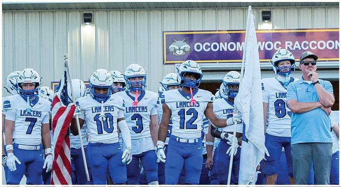 A group of people wearing helmets and sports gear stand in front of a building with a sign that reads “Oconomowoc Raccoons." Description generated by AI