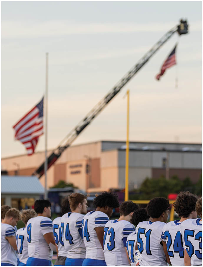 A group of people wearing white and blue sports uniforms stand in a line facing away with two American flags flying at half-mast in the background. Description generated by AI