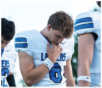 A person wearing a white and blue sports uniform is looking downward with another person partially visible beside them. Description generated by AI