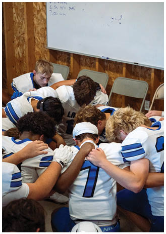A group of people wearing sports uniforms are huddled closely together with heads bowed and arms around each other in a room with chairs and a whiteboard. Description generated by AI