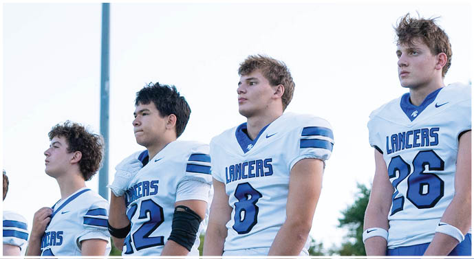 Four people wearing white football jerseys with “Lancers" written on them stand side by side outdoors, looking forward with serious expressions. Description generated by AI