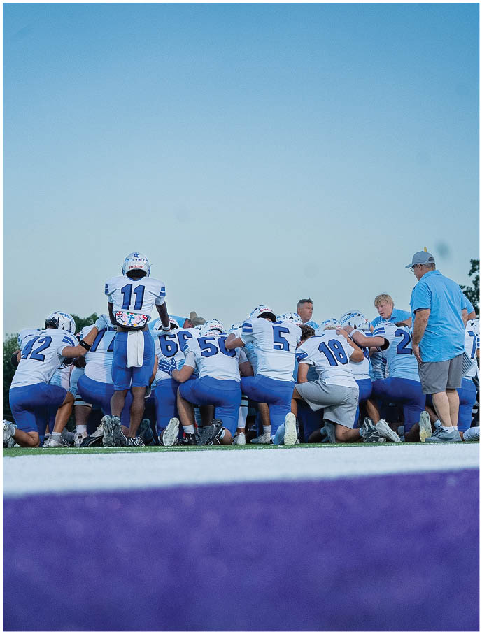 A group of people wearing football uniforms are kneeling together on a field under a clear sky while one person stands among them. Description generated by AI