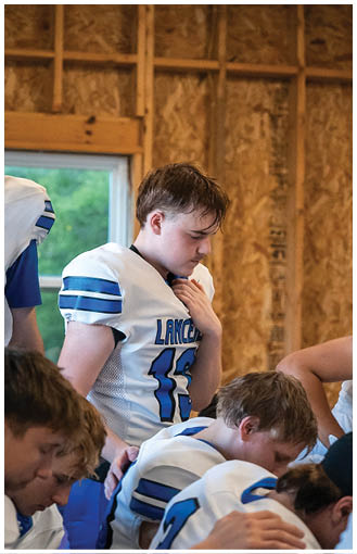 A person wearing a sports jersey with the word “Lancers" is looking down with a hand placed near their chest in an indoor setting with wooden walls. Description generated by AI