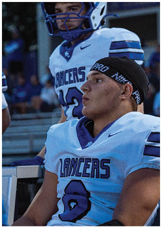 Two people wearing white football uniforms with “Lancers" written on the front are sitting on a bench, one wearing a helmet and the other wearing a black headband. Description generated by AI