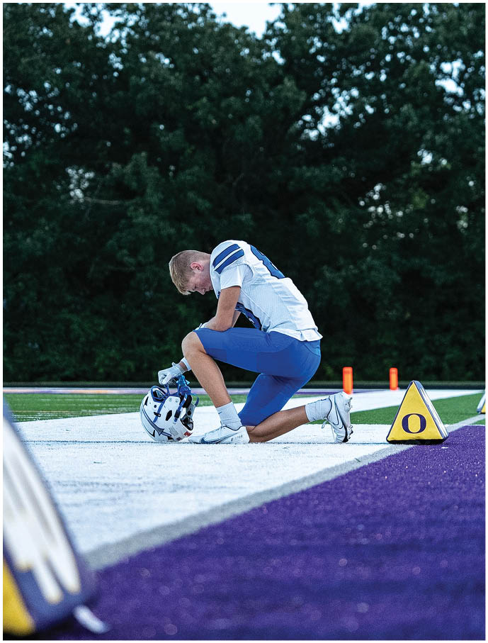 A person in a football uniform kneels on one knee on a field, holding a helmet on the ground near a yellow marker with the letter O. Description generated by AI