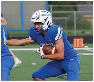 People wearing blue and white football uniforms are engaged in a game on a field with trees and a fence in the background. Description generated by AI