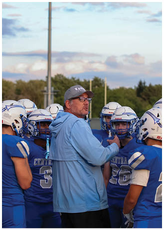 A person wearing glasses, a cap, and a light blue jacket is speaking to a group of people in blue football uniforms and helmets on a field with goalposts and trees in the background. Description generated by AI