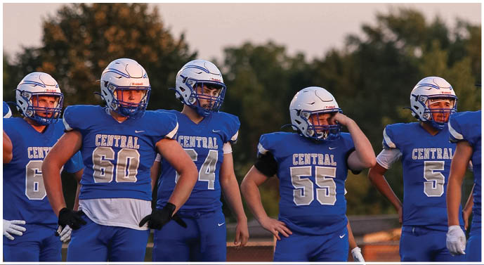 Five people wearing blue football uniforms and helmets stand outdoors with trees in the background during sunset. Description generated by AI