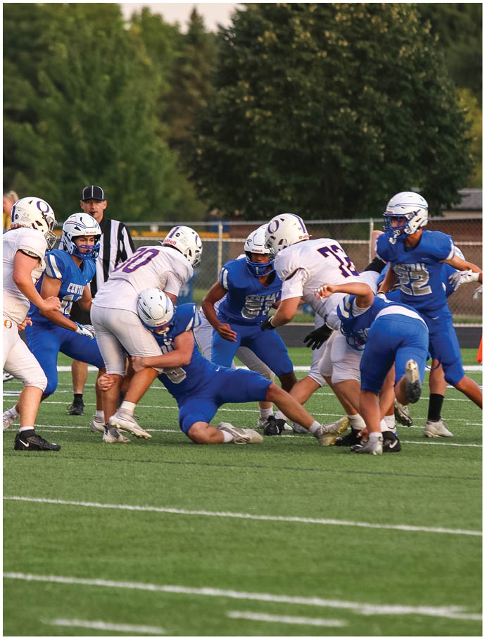 A group of people in blue and white football uniforms are engaged in a tackle on a green field with trees and a referee in the background. Description generated by AI