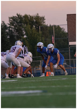 Two people in blue football uniforms are crouched in a ready position facing three people in white football uniforms on a grassy field during a game. Description generated by AI