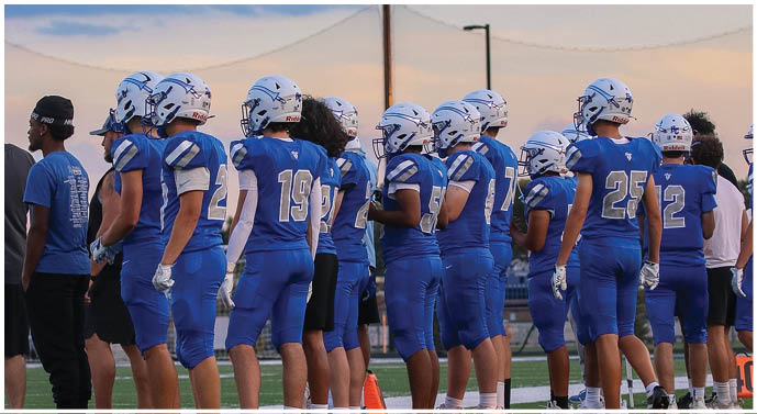 A group of people wearing blue football uniforms and helmets stand together on a field during sunset. Description generated by AI