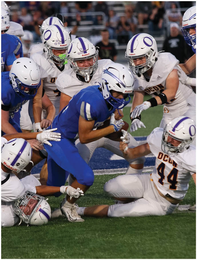 A group of people wearing football helmets and uniforms are engaged in a play on a football field with one person in blue running while others in white attempt to tackle. Description generated by AI