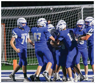 A group of people wearing blue football uniforms and white helmets are gathered near a goalpost on a field at night. Description generated by AI