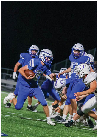 A group of people wearing blue football uniforms and helmets are actively playing on a field at night, with one person holding a football and running. Description generated by AI