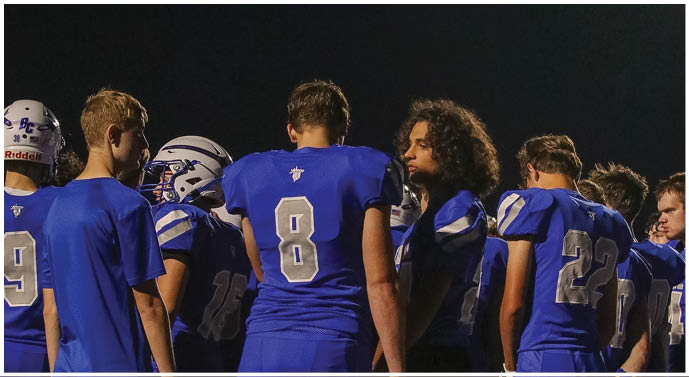 A group of people wearing blue sports jerseys are gathered closely together under a dark sky, with one person wearing a helmet. Description generated by AI