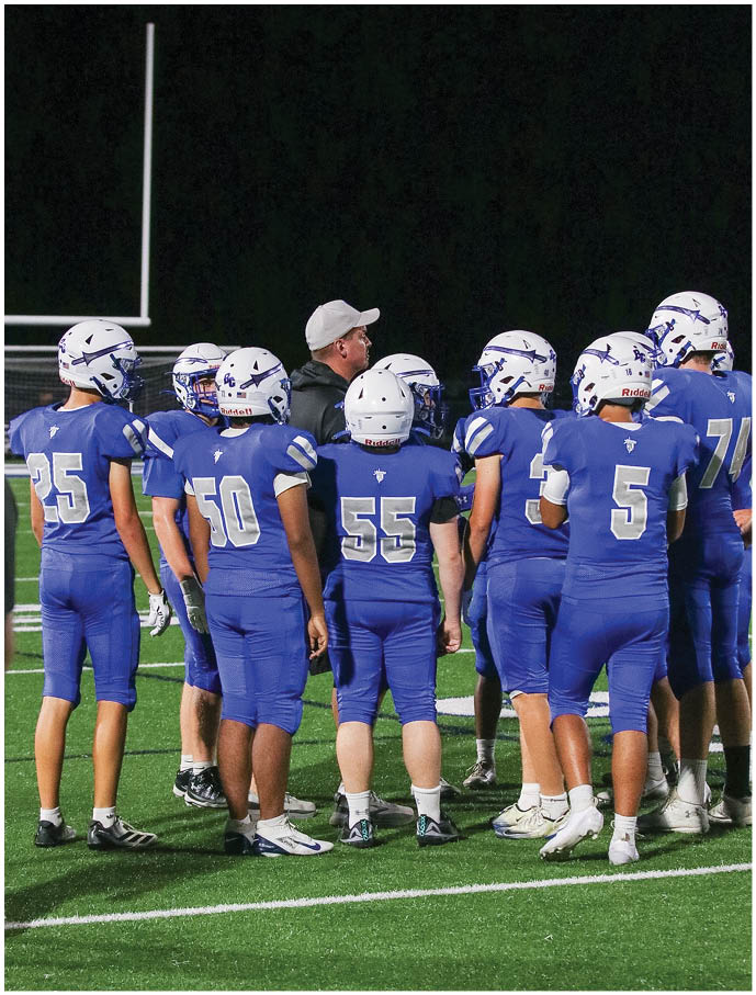 A group of people wearing blue football uniforms and helmets stand on a field at night while one person in a gray cap appears to be giving instructions. Description generated by AI
