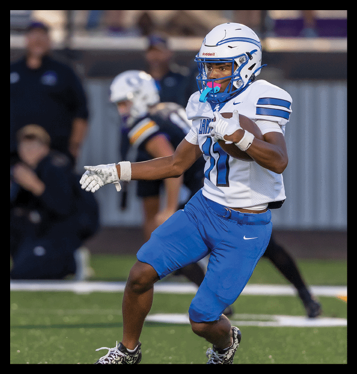 A person wearing a white and blue football uniform with a helmet and gloves is running on a field while holding a football. Description generated by AI