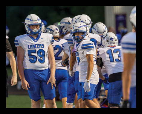 A group of people wearing white and blue football uniforms with helmets are standing together on a field near the sideline. Description generated by AI
