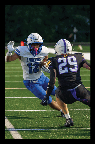 A person wearing a black and purple football uniform with the number 29 is in a defensive stance facing another person in a blue and white uniform on a green football field. Description generated by AI