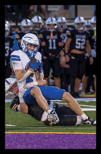 A person in a white and blue football uniform is being tackled by another person in a black football uniform on a field near the end zone with other players standing in the background. Description generated by AI