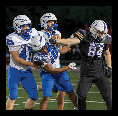 Three people in football uniforms are engaged in a play on a field with artificial turf and yard lines visible. Description generated by AI
