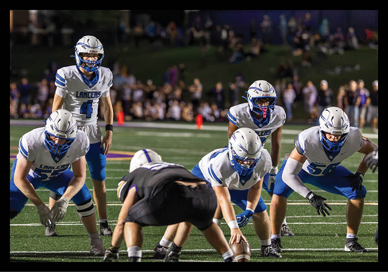 Several people in white and blue football uniforms are positioned on a field during a nighttime game with a crowd in the background. Description generated by AI