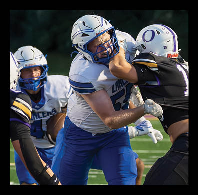 Two football players in white and blue uniforms are engaged in a physical play against players in black uniforms on a green field. Description generated by AI