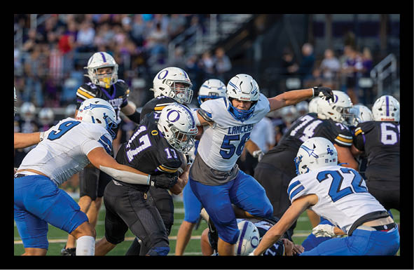 Players in blue and white and black and white football uniforms are actively engaged in a play on a football field with a crowd in the background. Description generated by AI