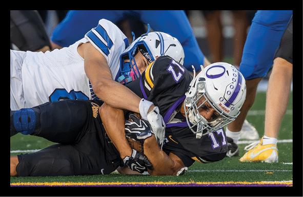 Two people in football uniforms are on the ground with one tackling the other during a game on a grassy field. Description generated by AI