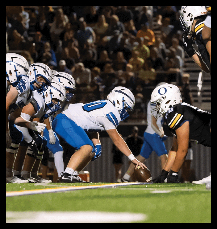 A group of football players in white helmets and blue uniforms are lined up on the field preparing for a play under stadium lights with a crowd in the background. Description generated by AI