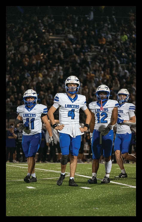 Four people in blue and white football uniforms with helmets walk together on a field with a crowd in the background. Description generated by AI
