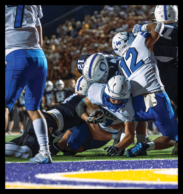 Several people in football uniforms are engaged in a tackle on a field during a game with a crowd in the background. Description generated by AI