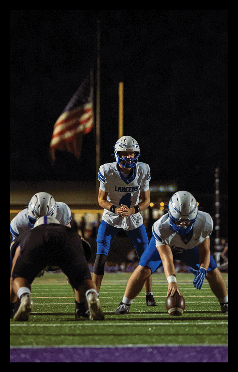 Three people in football uniforms are positioned on a field at night with one person holding a football and another standing behind them, ready to receive the ball, with an American flag and goalpost visible in the background. Description generated by AI