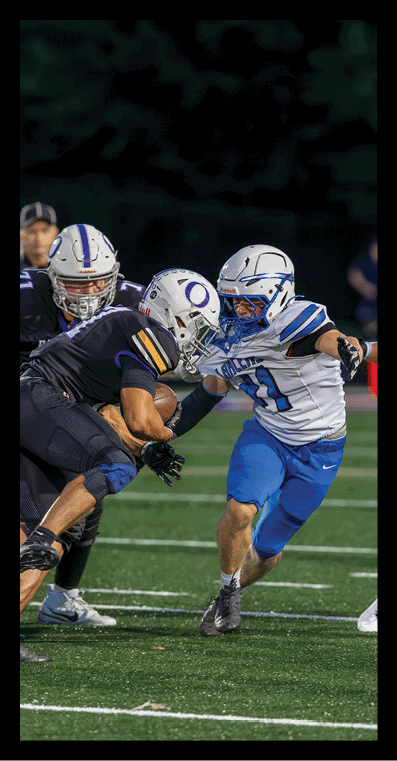 Two people in football uniforms are engaged in a tackle on a field during a game with a referee and other players visible in the background. Description generated by AI