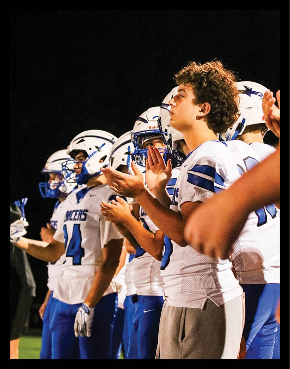 A person with curly hair is standing in front of several white helmets with blue star logos against a dark background. Description generated by AI