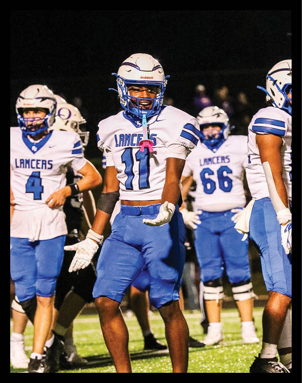 A person wearing a white football uniform with blue accents and a helmet is on a field at night with other similarly dressed people in the background. Description generated by AI